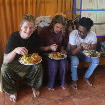 Trois personnes sont assises sur un banc, mangeant dans des assiettes de nourriture, avec un rideau jaune et des conteneurs de stockage à l'arrière-plan, dans une pièce au sol carrelé marron.