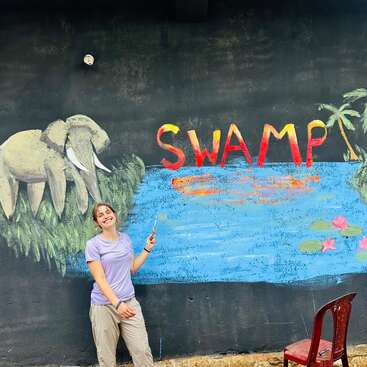 Eine junge Frau lächelt, während sie einen Pinsel in der Hand hält und vor einem bunten Wandbild steht, auf dem ein Elefant, ein Sumpf, eine Palme, Seerosen und das Wort „SWAMP“ zu sehen sind.