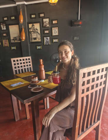 Une femme est assise à une table en bois dans un café chaleureux, un verre à la main. Les murs sombres sont ornés de photos encadrées et d'objets vintage.