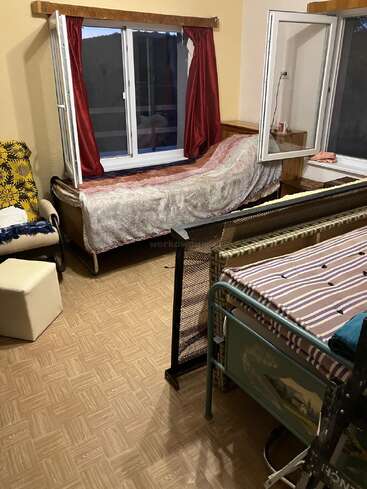 This cozy bedroom has two single beds, an armchair with a patterned blanket, red curtains, open windows, a nightstand, and parquet-style flooring. Warm atmosphere.