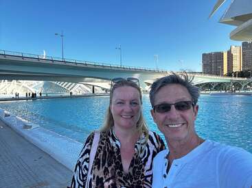 A smiling couple takes a selfie by a bright blue pool under a modern bridge, with clear skies and city buildings visible in the background.