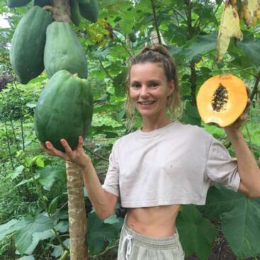 Une femme souriante se tient dans un jardin luxuriant, tenant une grande papaye verte sur l'arbre et une papaye mûre coupée, montrant ses graines.