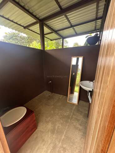 Il s'agit d'une salle de bains rustique, en plein air, avec des toilettes à compostage, un lavabo, un miroir et des murs partiels. Le toit est en métal et la verdure est visible à l'extérieur.