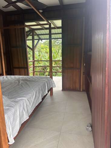 Cette image montre une chambre simple avec des murs en bois, un lit défait, un sol carrelé, des portes en bois ouvertes, une vue sur une végétation luxuriante et un ventilateur au plafond.