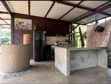 Cette image montre une cuisine rustique en plein air avec un îlot en béton, un réfrigérateur, une cuisinière, un filtre à eau, une tapisserie colorée, une œuvre d'art représentant une tortue et une végétation luxuriante à l'extérieur.