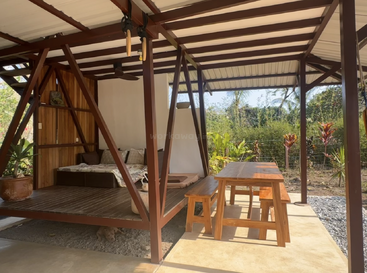 Cette image montre une structure en bois en plein air avec un lit ombragé et une table à manger en bois, des bancs, des plantes et de la verdure luxuriante en arrière-plan. Ambiance relaxante.