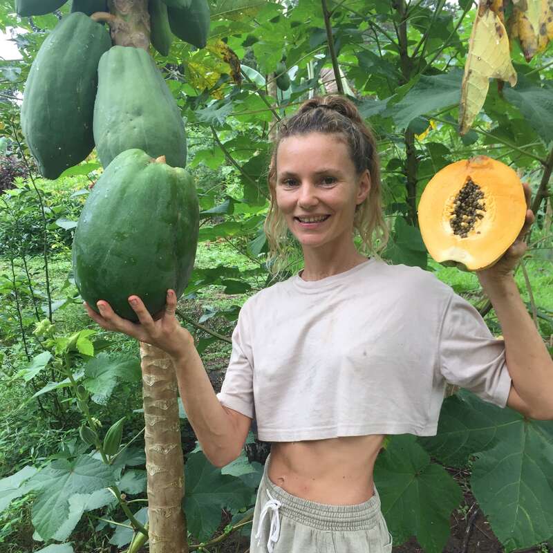 Une femme souriante se tient dans un jardin luxuriant, tenant une grande papaye verte sur l'arbre et une papaye mûre coupée, montrant ses graines.