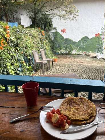 Ein Frühstück mit Omelett, Bananen und Papaya steht auf einem Holztisch im Freien. In der Nähe stehen eine rote Tasse, Gartenstühle, üppiges Grün und ein Wandgemälde.