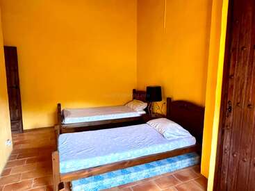 Esta imagen muestra una habitación sencilla con dos camas individuales de madera, paredes amarillas, suelo de baldosas, una lámpara negra, una decoración minimalista y un armario de madera.