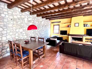 Esta acogedora sala de estar-comedor cuenta con paredes de piedra, vigas de madera a la vista, una chimenea amarilla, suelos de baldosas, muebles rústicos de madera, una lámpara colgante roja y cómodos asientos.