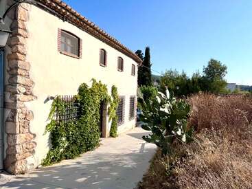 Una casa rústica de piedra con hiedra trepando por sus paredes, pequeñas ventanas enrejadas y techo de tejas se encuentra junto a un sendero, rodeada de hierba seca y cactus.