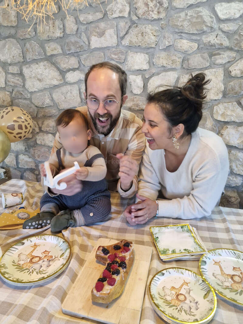 Una familia feliz celebra el primer cumpleaños de un niño. La mesa tiene platos, servilletas y globos con motivos de animales, y una tarta con forma del número uno decorada con bayas.