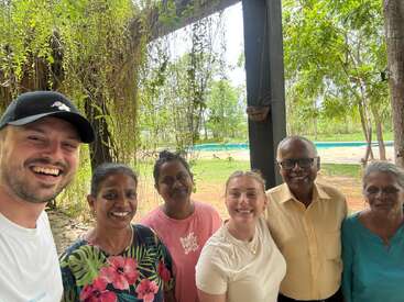 Eine Gruppe von sechs lächelnden Menschen posiert gemeinsam im Freien, umgeben von Grün und Bäumen. Die Atmosphäre ist fröhlich und entspannt, mit einer sonnigen, natürlichen Kulisse im Hintergrund.