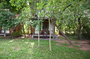 Eine urige Holzhütte, umgeben von üppigen grünen Bäumen und Gras, mit einer Veranda und entspannenden Stühlen, die eine friedliche und natürliche Rückzugsatmosphäre schaffen.