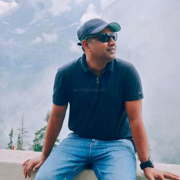 Ein Mann mit Sonnenbrille, Mütze, dunklem Poloshirt und Jeans sitzt auf einem Felsvorsprung vor einer nebligen Bergkulisse und schaut nachdenklich in die Ferne.