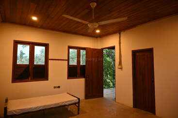 Dieses Bild zeigt ein einfaches Zimmer mit einem Einzelbett, Holztüren und -fenstern, einem Deckenventilator und einer Öffnung zu einem grünen Außenbereich.