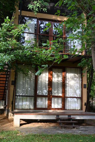 Ein modernes zweistöckiges Gebäude mit großen Glasfenstern und Holztüren, umgeben von üppigen grünen Bäumen, die sich harmonisch in die natürliche Umgebung einfügen. Eine Treppe führt zum Eingang hinauf.