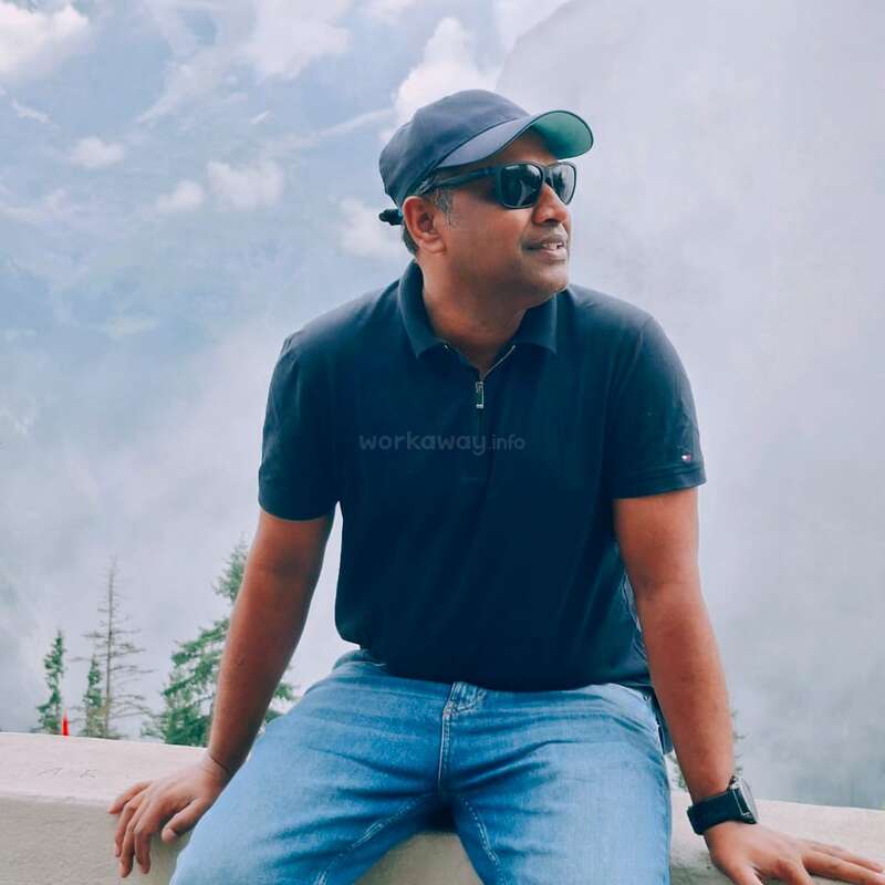 Ein Mann mit Sonnenbrille, Mütze, dunklem Poloshirt und Jeans sitzt auf einem Felsvorsprung vor einer nebligen Bergkulisse und schaut nachdenklich in die Ferne.