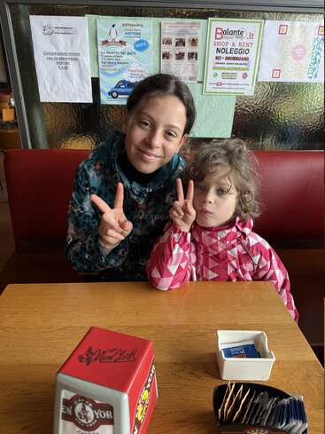Two children sit at a wooden table in a cafe, smiling and making peace signs. Behind them, colorful posters are displayed on a glass partition wall.
