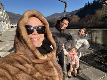 The image shows a family of four posing for a selfie on a sunny day, with mountains and trees in the background.