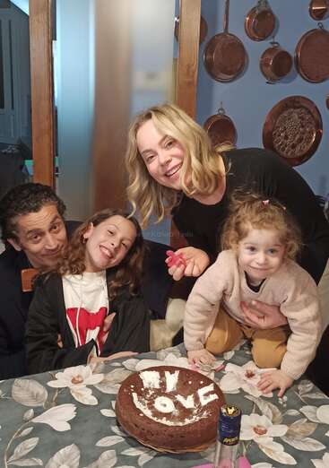 The image depicts a family of four gathered around a table with a cake featuring the words \"MY LOVE\" in icing, set against a blue wall with copper pans hanging on it.