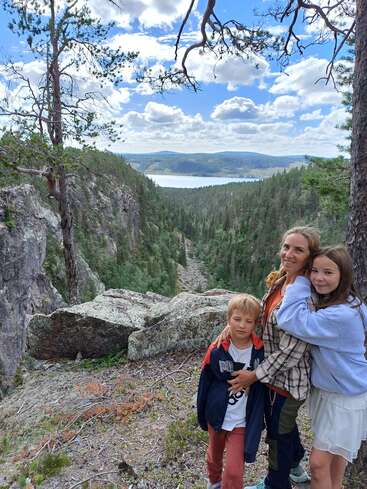 The image depicts a woman and two children standing on a rocky hillside, surrounded by trees and a lake in the distance, with a blue sky and white clouds above.