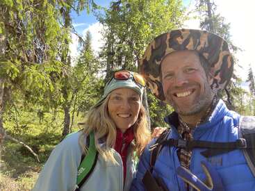 The image depicts a man and woman posing for a photo in a wooded area, both wearing hats and jackets, with the man sporting a backpack.