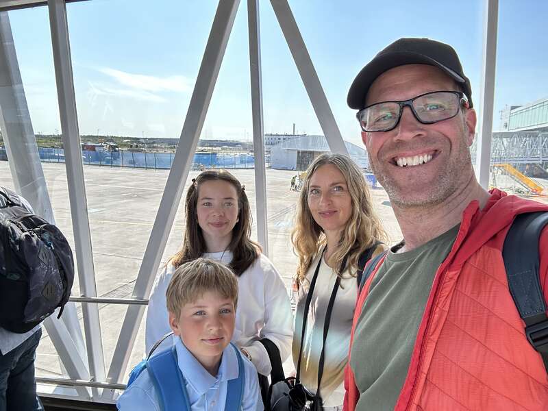 A smiling family of four poses for a selfie at an airport boarding bridge, with sunny weather outside and luggage on their backs, ready for travel.