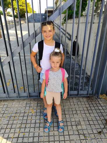 Zwei junge Mädchen mit Rucksäcken stehen vor einem eingezäunten Gelände. Das ältere Mädchen lächelt, während das jüngere ernst dreinschaut. Sie scheinen bereit für die Schule zu sein.
