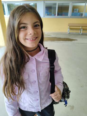 Una joven sonriente con el pelo largo y castaño está fuera, lleva una chaqueta morada clara y una gargantilla negra, y sujeta una mochila. Está en el colegio.