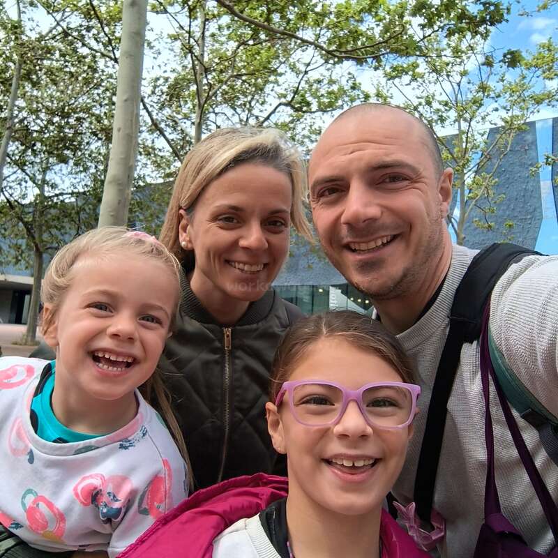 Eine glückliche vierköpfige Familie, zwei Erwachsene und zwei Kinder, lächelt an einem hellen Tag im Freien, umgeben von grünen Bäumen und modernen architektonischen Gebäuden im Hintergrund.