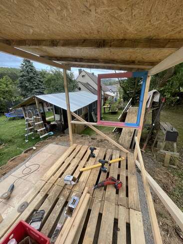 Une structure en bois en cours de construction, avec des outils tels que des perceuses, un niveau et du matériel sur des palettes en bois, en plein air dans une arrière-cour avec des arbres et des bâtiments.