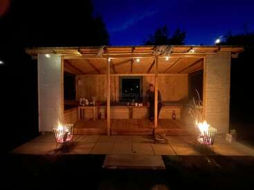 A cozy wooden cabin is illuminated at night by warm fire pits on both sides, with a person sitting inside, creating a peaceful and inviting atmosphere.