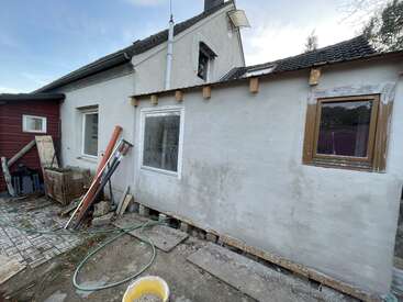 The image shows a house under renovation with construction materials and debris scattered around. There are exposed beams, unfinished walls, and some tools lying on the ground.