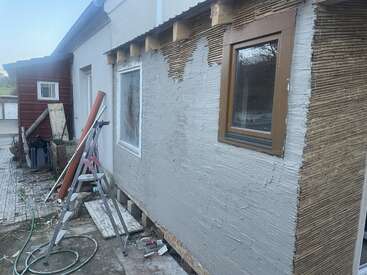 A partially plastered house wall is under renovation. Construction materials, tools, and a ladder are present. The wall shows visible laths and fresh stucco application.