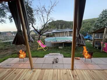 A cozy backyard scene with two fire pits, lounge chairs, an above-ground pool, a wooden deck, a tree, and rustic outdoor structures, surrounded by greenery.