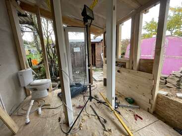 This image shows a small wooden building under construction with a toilet, tools, and construction materials scattered. There’s a work light, cables, and exposed framing visible.