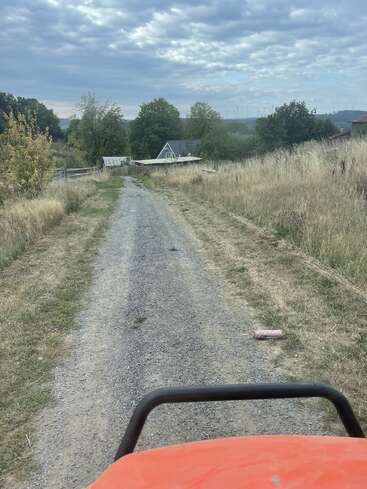 A gravel path leads to a house surrounded by trees and dry grass, under a cloudy sky. An orange vehicle's front is visible, and a paint roller lies discarded.