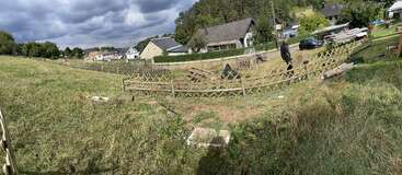 A rural area with grassy fields, a partially collapsed wooden fence, scattered debris, one person standing, houses in the background, and cloudy skies overhead.