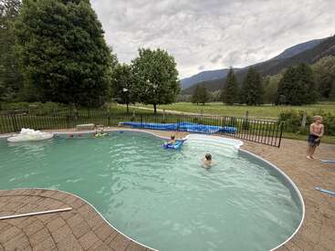 Tres niños disfrutan de una piscina rodeada de árboles y montañas. En el agua flotan juguetes de piscina. Cielo nublado, un tranquilo paisaje rural y un patio vallado.