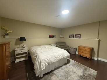 Este acogedor dormitorio cuenta con una cama blanca, muebles de madera, paredes en tonos beige, una manta suave, cuadros enmarcados, un sillón de lectura, una lámpara y una decoración discreta y acogedora.