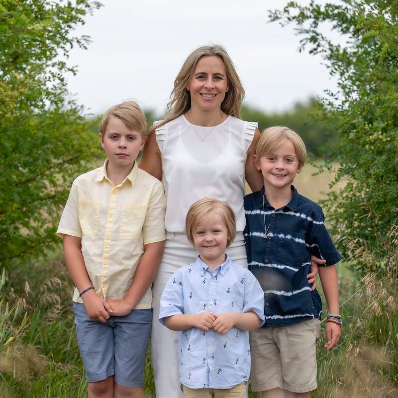 Una mujer está al aire libre con tres niños pequeños; todos sonríen, excepto uno. Están rodeados de arbustos verdes y césped, disfrutando juntos de un momento en familia.