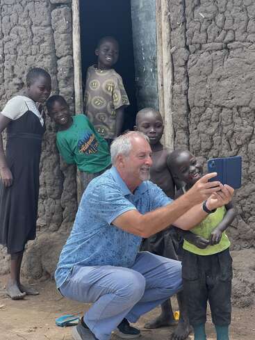 Eine Gruppe lächelnder Kinder und ein Mann machen gemeinsam ein Selfie vor einem rustikalen Haus mit Lehmwänden und fangen einen fröhlichen und herzerwärmenden Moment der Verbundenheit ein.