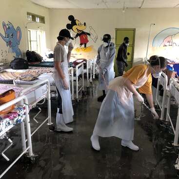 Das Bild zeigt eine Gruppe von Menschen, die ein Krankenhauszimmer mit weißen Betten und Cartoonfiguren an den Wänden reinigen.