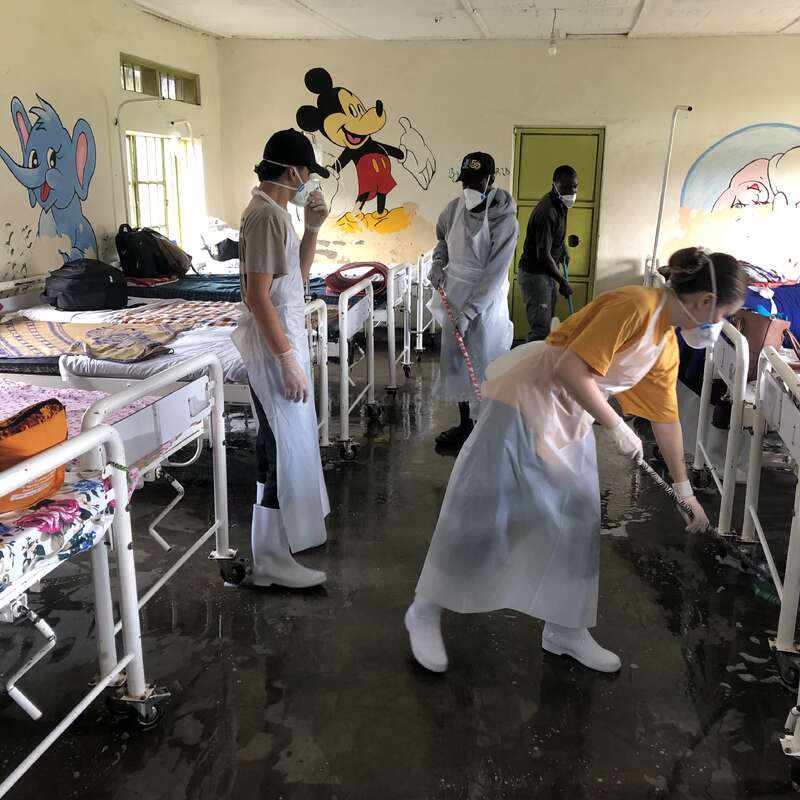 Das Bild zeigt eine Gruppe von Menschen, die ein Krankenhauszimmer mit weißen Betten und Cartoonfiguren an den Wänden reinigen.