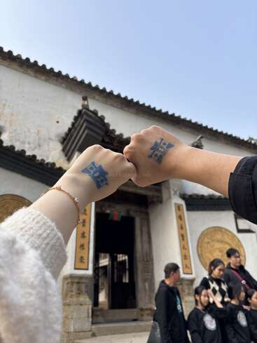Duas pessoas mostram tatuagens azuis iguais em suas mãos, formando punhos juntos em frente a um edifício tradicional chinês, com um grupo de pessoas ao fundo.