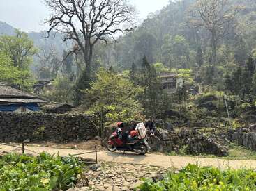 Uma paisagem rural exuberante com folhagem verde, paredes de pedra e casas espalhadas. Uma pessoa dirige uma motocicleta vermelha enquanto outra se senta em uma rocha próxima. Cena tranquila.