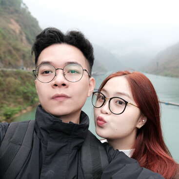 Ein junges Paar, das eine Brille trägt, macht ein Selfie im Freien an einem nebligen Fluss, umgeben von üppigen grünen Bergen und einer nebligen, ruhigen Naturkulisse im Hintergrund.