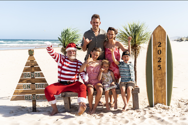 Une famille joyeuse pose avec le Père Noël sur une plage ensoleillée. Une planche de surf marquée « 2025 », une pancarte en forme d'arbre de Noël et l'ambiance festive créent une scène estivale unique.