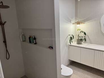Cette salle de bains moderne comprend une douche à l'italienne avec pomme de douche à effet pluie, une étagère intégrée pour les articles de toilette, un meuble-lavabo suspendu, une plante d'intérieur, un luminaire suspendu et une décoration minimaliste blanche.